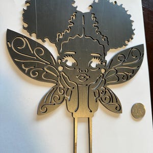 May include: A metal fairy cake topper with a girl's face, afro puffs, and ornate wings. The fairy's hands are resting on her chin. The topper is silver in color and has two prongs at the bottom for insertion into a cake.