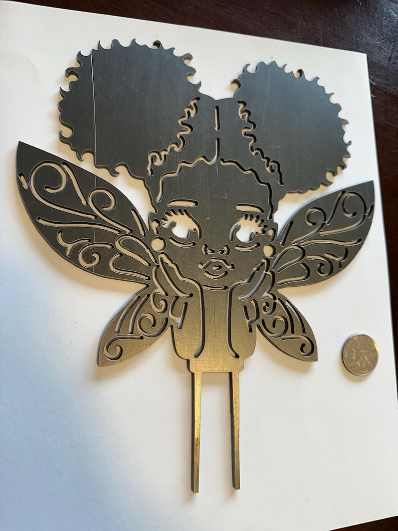 May include: A metal fairy cake topper with a girl's face, afro puffs, and ornate wings. The fairy's hands are resting on her chin. The topper is silver in color and has two prongs at the bottom for insertion into a cake.