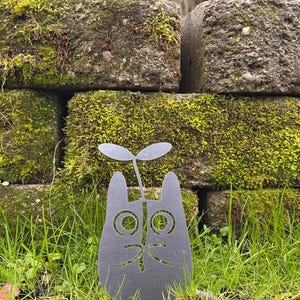 May include: A dark gray metal cat-shaped garden stake with a sprout growing from its head. The cat has cut-out eyes, nose, and mouth. The stake is in a grassy area in front of a moss-covered stone wall.