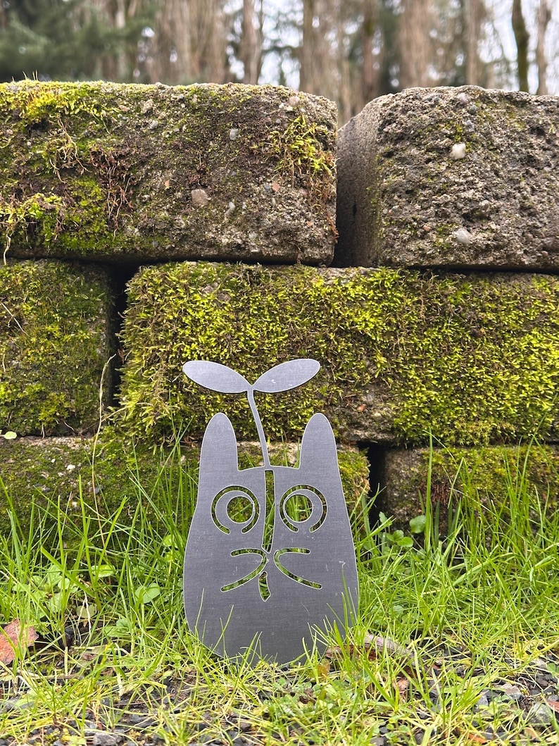 May include: A dark gray metal cat-shaped garden stake with a sprout growing from its head. The cat has cut-out eyes, nose, and mouth. The stake is in a grassy area in front of a moss-covered stone wall.