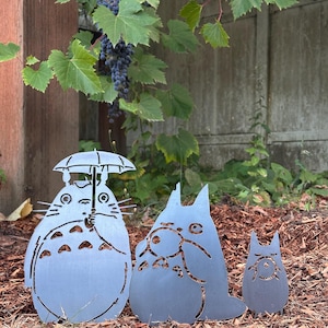 May include: Three metal garden art figures in the shape of characters from a popular animated film. The largest figure holds an umbrella. The figures are gray and stand on a bed of brown mulch, with green foliage and a concrete wall in the background.