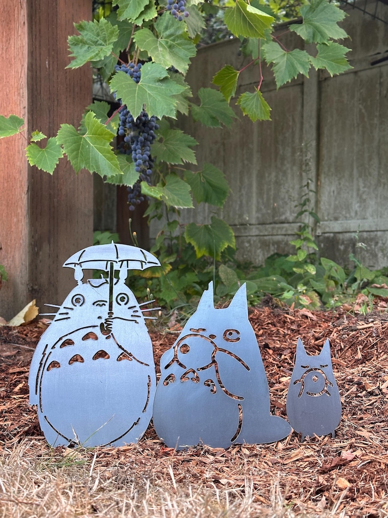 May include: Three metal garden art figures in the shape of characters from a popular animated film. The largest figure holds an umbrella. The figures are gray and stand on a bed of brown mulch, with green foliage and a concrete wall in the background.