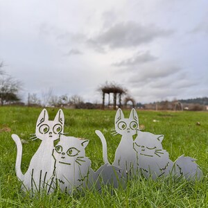May include: Four metal cat yard art pieces are displayed in a grassy field under an overcast sky. The cats are cut from a silver-colored metal and have large eyes and long tails. The cats are in pairs, with one cat cuddling the other.