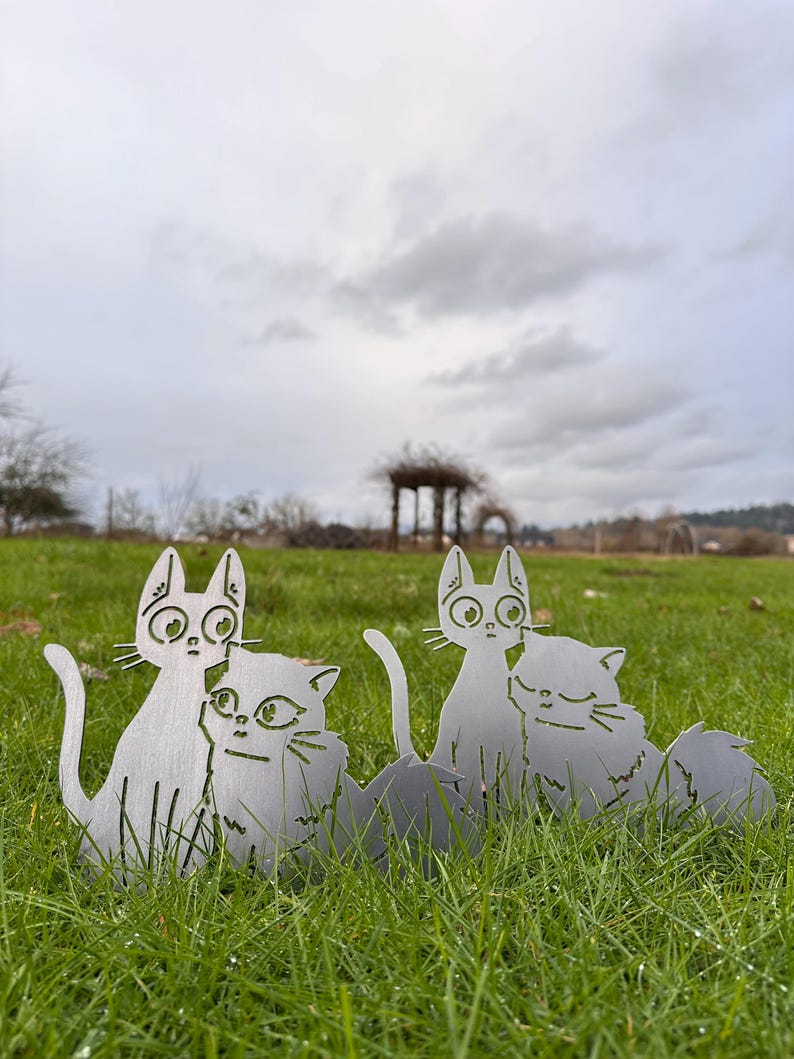 May include: Four metal cat yard art pieces are displayed in a grassy field under an overcast sky. The cats are cut from a silver-colored metal and have large eyes and long tails. The cats are in pairs, with one cat cuddling the other.