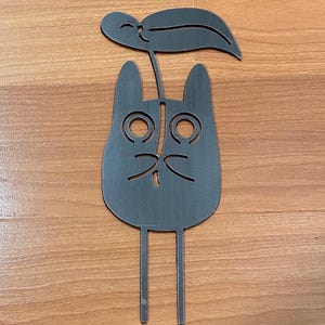 May include: A dark gray metal cat-shaped garden stake with a leaf on top. The cat has large, round eyes and a small, curved mouth. The stake has two thin legs for insertion into the ground. The design is simple and whimsical.