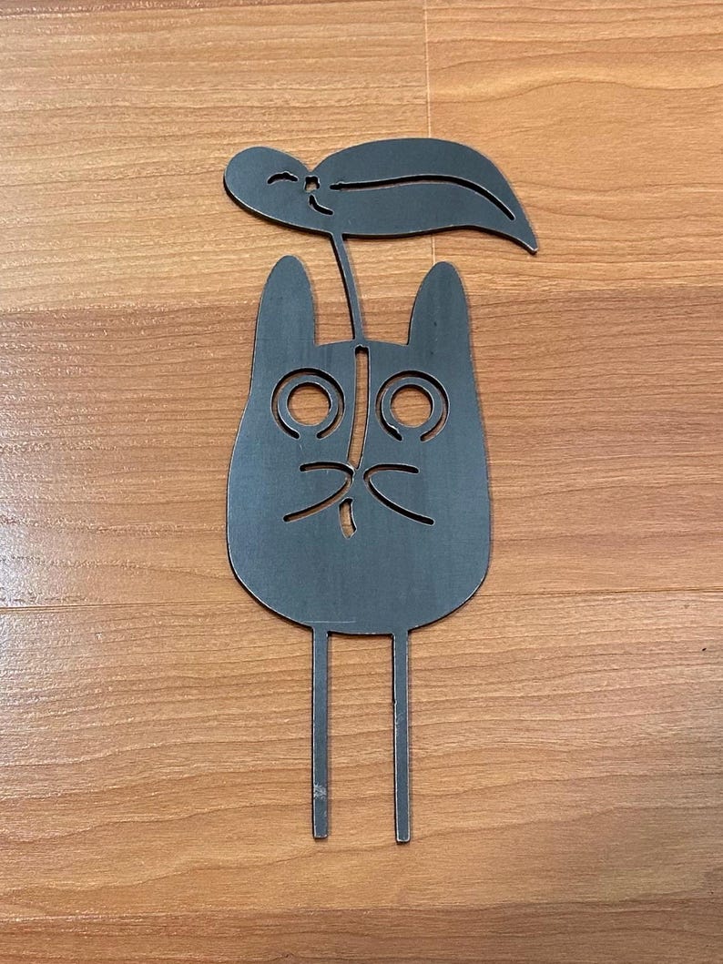 May include: A dark gray metal cat-shaped garden stake with a leaf on top. The cat has large, round eyes and a small, curved mouth. The stake has two thin legs for insertion into the ground. The design is simple and whimsical.