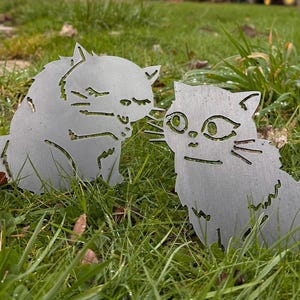 May include: Two metal cat yard art pieces are displayed in a grassy area. The cats are cut from a silver-colored metal and have detailed features. One cat is facing the other, creating a charming outdoor decoration.