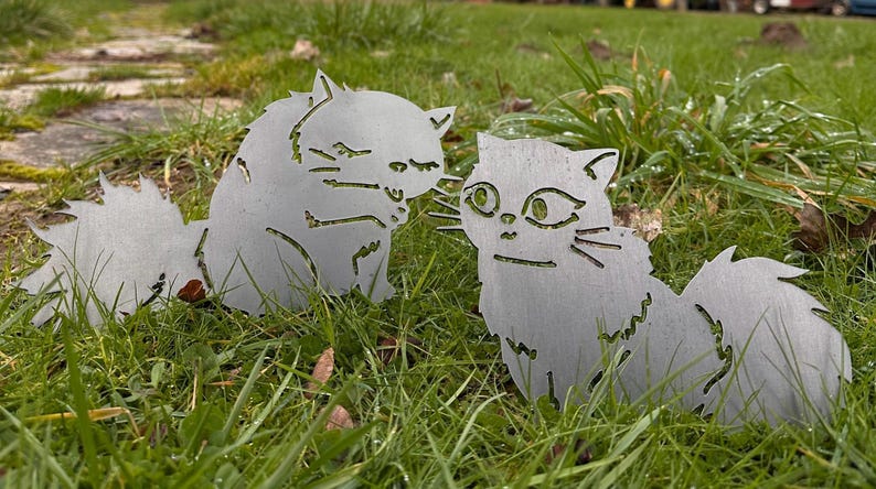 May include: Two metal cat yard art pieces are displayed in a grassy area. The cats are cut from a silver-colored metal and have detailed features. One cat is facing the other, creating a charming outdoor decoration.