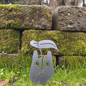 May include: A gray metal cat-shaped garden stake with a leaf design. The cat has large, round eyes and a smiling mouth. The stake is placed in green grass in front of a moss-covered brick wall.