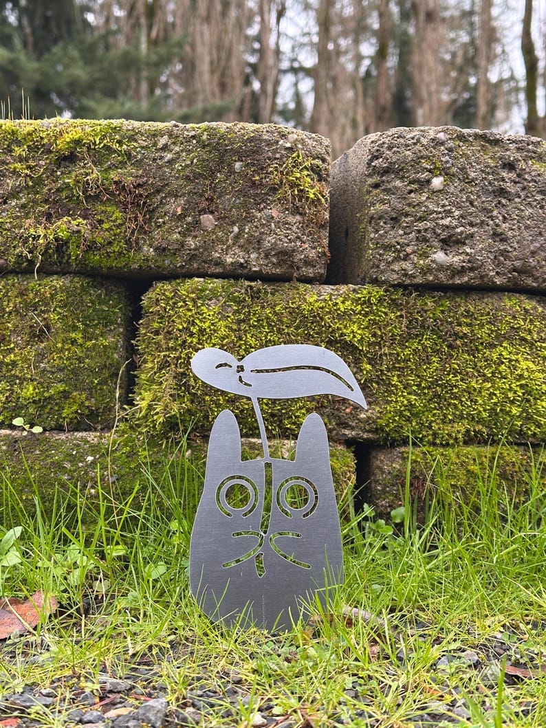 May include: A gray metal cat-shaped garden stake with a leaf design. The cat has large, round eyes and a smiling mouth. The stake is placed in green grass in front of a moss-covered brick wall.
