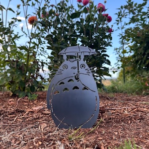 May include: A dark gray metal garden art piece depicting a cartoon character holding an umbrella. The silhouette is set in a garden bed with brown mulch, green grass, and colorful flowers in the background.