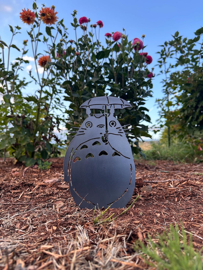 May include: A dark gray metal garden art piece depicting a cartoon character holding an umbrella. The silhouette is set in a garden bed with brown mulch, green grass, and colorful flowers in the background.