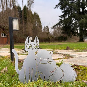 May include: A metal cat sculpture featuring two cats in an embrace, set in a grassy outdoor environment. The cats are cut from a silver-colored metal, with details like eyes and whiskers. The sculpture is placed on a bed of green grass.