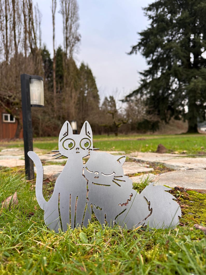 May include: A metal cat sculpture featuring two cats in an embrace, set in a grassy outdoor environment. The cats are cut from a silver-colored metal, with details like eyes and whiskers. The sculpture is placed on a bed of green grass.