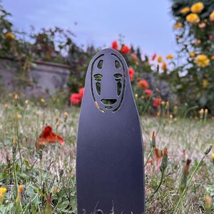 May include: A black metal cutout of the No-Face spirit from Spirited Away, standing in a grassy area with flowers. The cutout has a distinctive mask-like face with large eyes and a smiling mouth. The background features red and yellow flowers.
