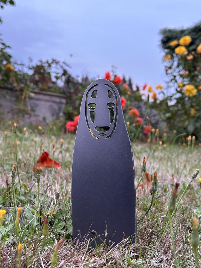 May include: A black metal cutout of the No-Face spirit from Spirited Away, standing in a grassy area with flowers. The cutout has a distinctive mask-like face with large eyes and a smiling mouth. The background features red and yellow flowers.