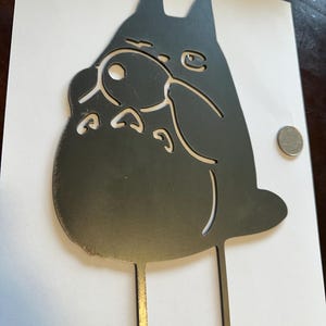 May include: A black metal silhouette of a cartoon character with large ears and a round belly. The character has cut-out details for eyes, nose, and mouth. Two thin metal rods extend from the bottom of the character.