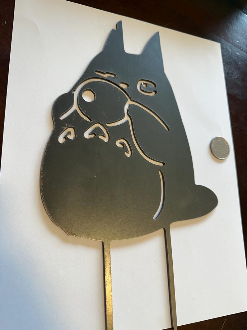 May include: A black metal silhouette of a cartoon character with large ears and a round belly. The character has cut-out details for eyes, nose, and mouth. Two thin metal rods extend from the bottom of the character.