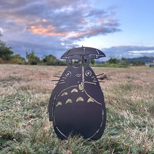 May include: A black metal silhouette of a Totoro character holding an umbrella. The artwork is placed in a grassy field with a cloudy sky in the background. The silhouette has cut-out details.