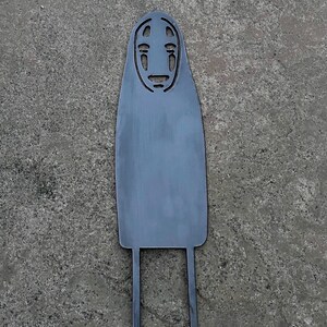 May include: A gray metal cutout of the character No-Face from the animated film Spirited Away. The cutout features the character's distinctive mask-like face and long, flowing body with two legs extending from the bottom.