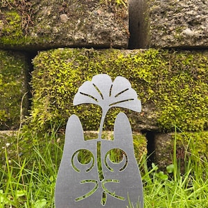May include: A dark gray metal cat silhouette with a cut-out face and a stylized leaf design above its head. The cat is set in a grassy area with a moss-covered brick wall in the background. The cat's features are cut out, creating a unique garden art piece.