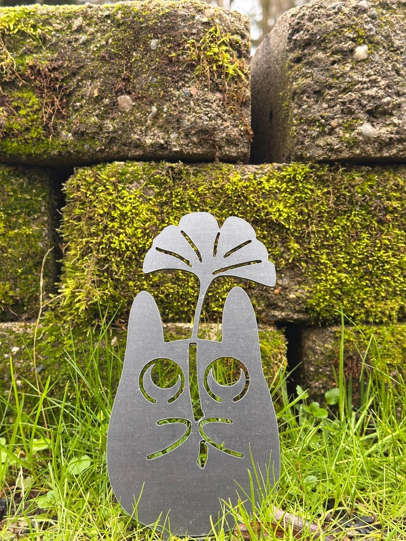 May include: A dark gray metal cat silhouette with a cut-out face and a stylized leaf design above its head. The cat is set in a grassy area with a moss-covered brick wall in the background. The cat's features are cut out, creating a unique garden art piece.