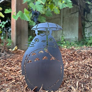 May include: A black metal garden sculpture of a cartoon character holding an umbrella. The sculpture has cut-out details and is placed in a garden bed with wood chips. The character has a large belly and a smiling face.