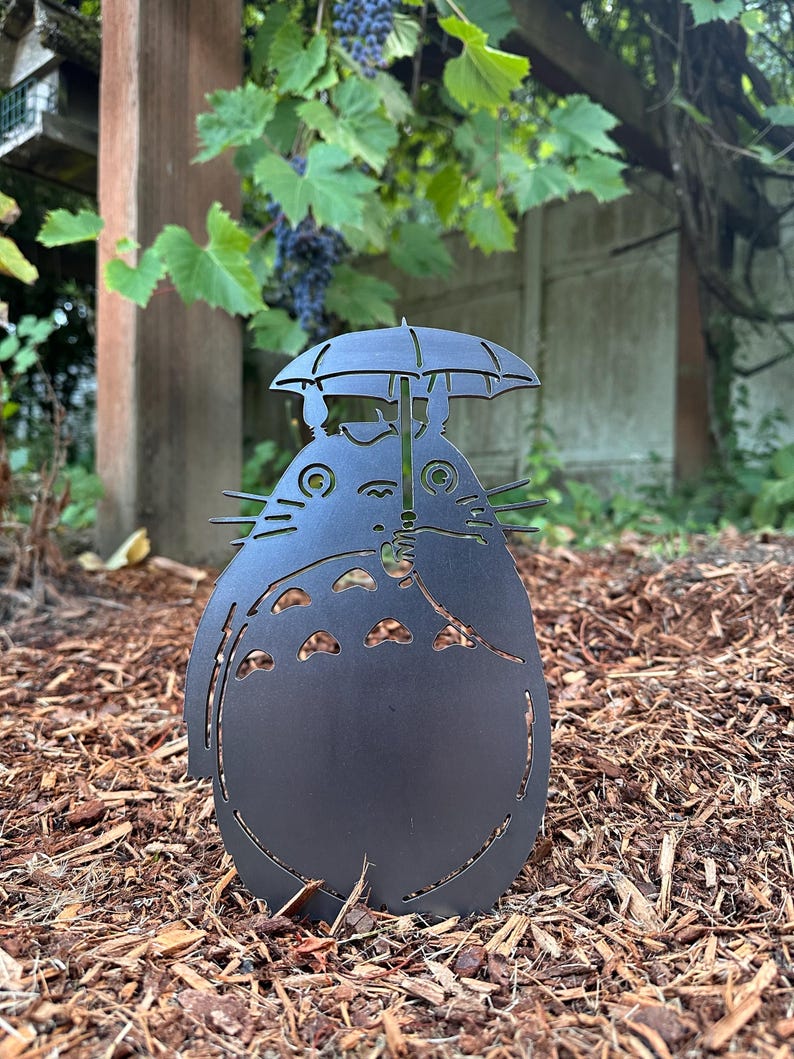 May include: A black metal garden sculpture of a cartoon character holding an umbrella. The sculpture has cut-out details and is placed in a garden bed with wood chips. The character has a large belly and a smiling face.