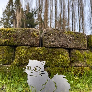 May include: A silver metal cat silhouette sits in green grass. The cat has a fluffy tail and is in front of a moss-covered brick wall. The background includes trees and a cloudy sky.