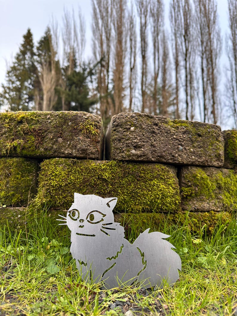 May include: A silver metal cat silhouette sits in green grass. The cat has a fluffy tail and is in front of a moss-covered brick wall. The background includes trees and a cloudy sky.