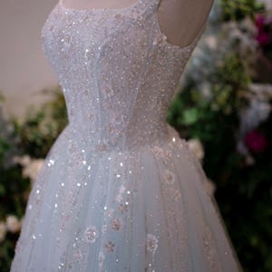 May include: A light blue formal gown on a mannequin. The dress features a square neckline, embellished with sequins and floral appliques. The tulle skirt is adorned with scattered sequins and floral details, creating a shimmering effect. The dress is designed for special occasions.