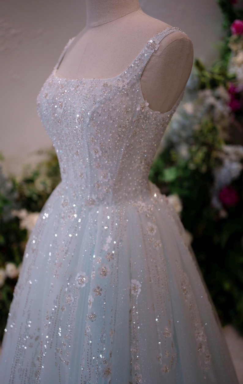 May include: A light blue formal gown on a mannequin. The dress features a square neckline, embellished with sequins and floral appliques. The tulle skirt is adorned with scattered sequins and floral details, creating a shimmering effect. The dress is designed for special occasions.
