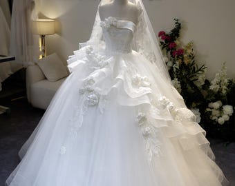 May include: A strapless white wedding gown with a full skirt and layered ruffles. The bodice is adorned with floral appliqués and the mannequin is topped with a gold crown and veil. The dress is displayed in a bridal shop.