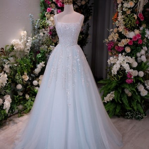 May include: A light blue, floor-length gown with a square neckline and a full skirt. The bodice is embellished with sequins and floral appliques. The dress is displayed on a mannequin in front of a floral archway, creating a romantic and elegant aesthetic.
