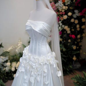 May include: A white strapless wedding gown featuring a structured corset bodice with draped fabric across the chest, a full skirt adorned with three-dimensional white floral appliqu&eacute;s, and a long sheer veil topped with a gold crown headpiece.