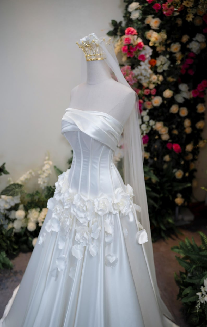 May include: A white strapless wedding gown featuring a structured corset bodice with draped fabric across the chest, a full skirt adorned with three-dimensional white floral appliqu&eacute;s, and a long sheer veil topped with a gold crown headpiece.