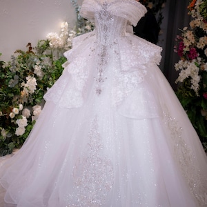 May include: A white ball gown featuring an off-the-shoulder neckline, a corset-style bodice with silver beadwork, and a voluminous skirt with layered lace appliques and glittery fabric. A small gold crown rests on the top of the mannequin.