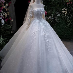 May include: A white bridal gown with a full skirt and long sleeves. The bodice and sleeves feature floral detailing. A sheer veil and a crown are also present. The dress is displayed on a mannequin against a floral backdrop.