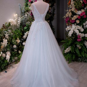 May include: A light blue wedding gown on a mannequin, featuring a V-neckline and a flowing tulle skirt. The dress is adorned with delicate floral embellishments and a lace-up back. The gown is displayed in front of a floral backdrop.