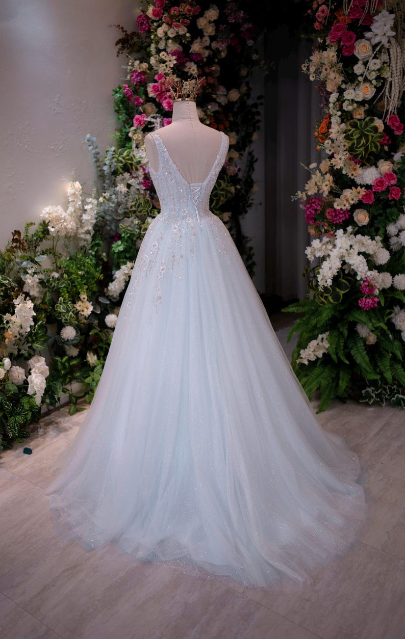 May include: A light blue wedding gown on a mannequin, featuring a V-neckline and a flowing tulle skirt. The dress is adorned with delicate floral embellishments and a lace-up back. The gown is displayed in front of a floral backdrop.