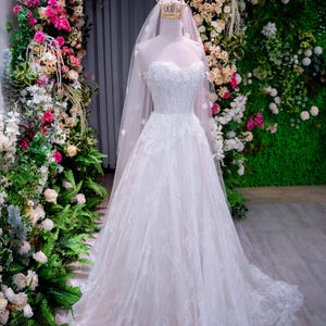May include: A strapless white wedding gown with intricate lace detailing. The dress features a full skirt and a long, flowing train. A sheer veil and a gold crown are on the mannequin. The background includes a floral arch with pink and white flowers.