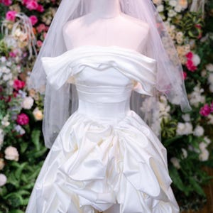 May include: A white satin wedding dress on a mannequin, with an off-the-shoulder neckline and a full skirt with ruffled details. A gold crown and veil are on the mannequin's head. The background is a floral arrangement.