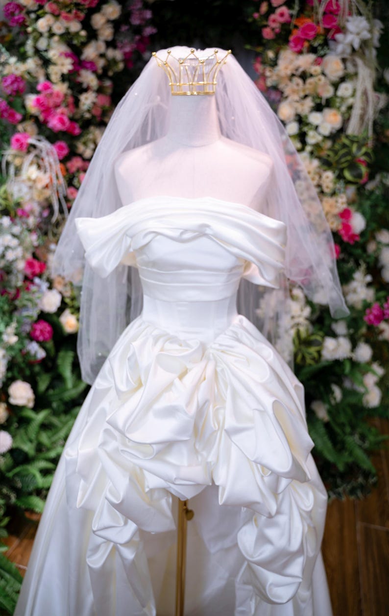 May include: A white satin wedding dress on a mannequin, with an off-the-shoulder neckline and a full skirt with ruffled details. A gold crown and veil are on the mannequin's head. The background is a floral arrangement.