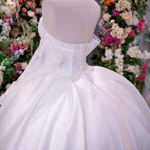 May include: A white strapless ball gown featuring a corset-style bodice with lace detailing, a lace-up back closure, and a voluminous satin skirt. The dress is displayed on a mannequin wearing a gold crown, showcasing a structured silhouette.