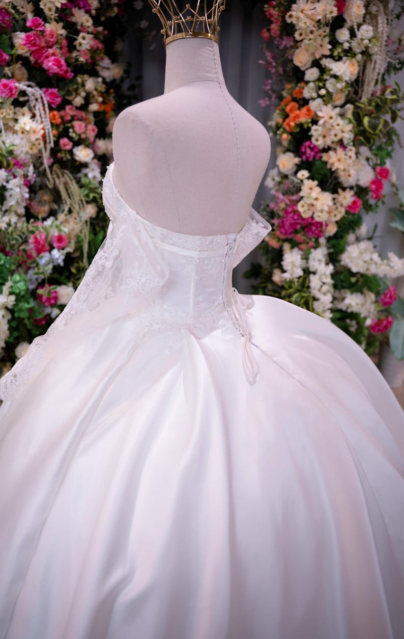 May include: A white strapless ball gown featuring a corset-style bodice with lace detailing, a lace-up back closure, and a voluminous satin skirt. The dress is displayed on a mannequin wearing a gold crown, showcasing a structured silhouette.