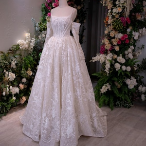 K&ouml;nnte beinhalten: Wei&szlig;es Brautkleid auf einer Schaufensterpuppe, mit quadratischem Ausschnitt, langen &Auml;rmeln und weitem Rock. Das Kleid ist mit aufwendiger Spitze und schimmernden Verzierungen versehen. Der Hintergrund umfasst einen Blumenbogen und Gr&uuml;n.