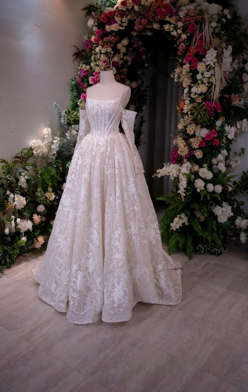 K&ouml;nnte beinhalten: Wei&szlig;es Brautkleid auf einer Schaufensterpuppe, mit quadratischem Ausschnitt, langen &Auml;rmeln und weitem Rock. Das Kleid ist mit aufwendiger Spitze und schimmernden Verzierungen versehen. Der Hintergrund umfasst einen Blumenbogen und Gr&uuml;n.