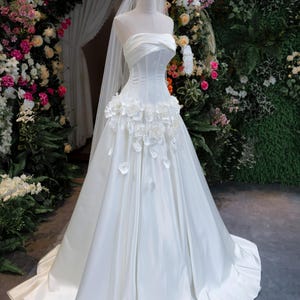 May include: A strapless white wedding dress featuring a structured bodice with horizontal pleats and a full-length skirt. The waistline is adorned with a cluster of white fabric flowers. A long, sheer white veil is attached to a gold-toned crown.