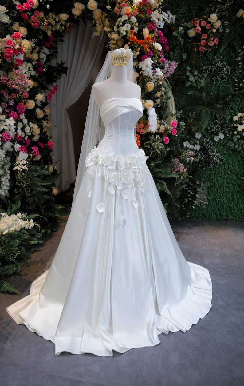 May include: A strapless white wedding dress featuring a structured bodice with horizontal pleats and a full-length skirt. The waistline is adorned with a cluster of white fabric flowers. A long, sheer white veil is attached to a gold-toned crown.