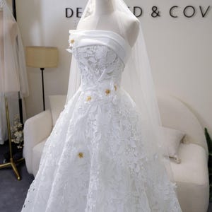 May include: A white bridal gown on a mannequin, with a strapless design and floral lace detailing. The dress has a full skirt and is decorated with small gold accents. A sheer veil is draped from the mannequin's head. The background includes the text "POMH DE D & COV".